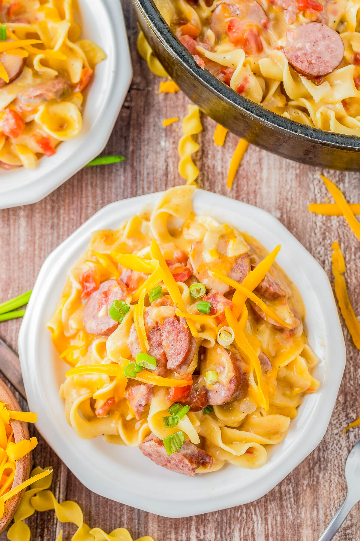 A plate of creamy pasta with sliced sausage, diced tomatoes, shredded cheddar cheese, and green onions, with a pan and another plate of the same dish in the background.