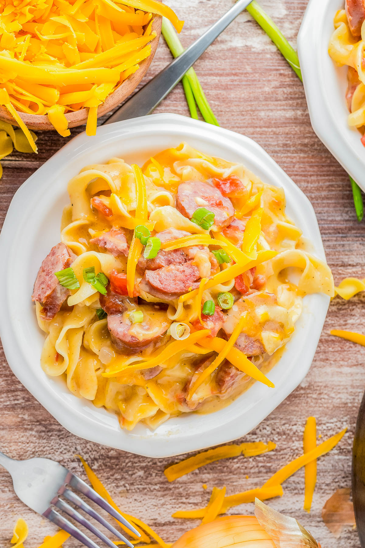 A plate of cheesy pasta with sliced sausage, garnished with chopped green onions and shredded cheese, on a wooden table.