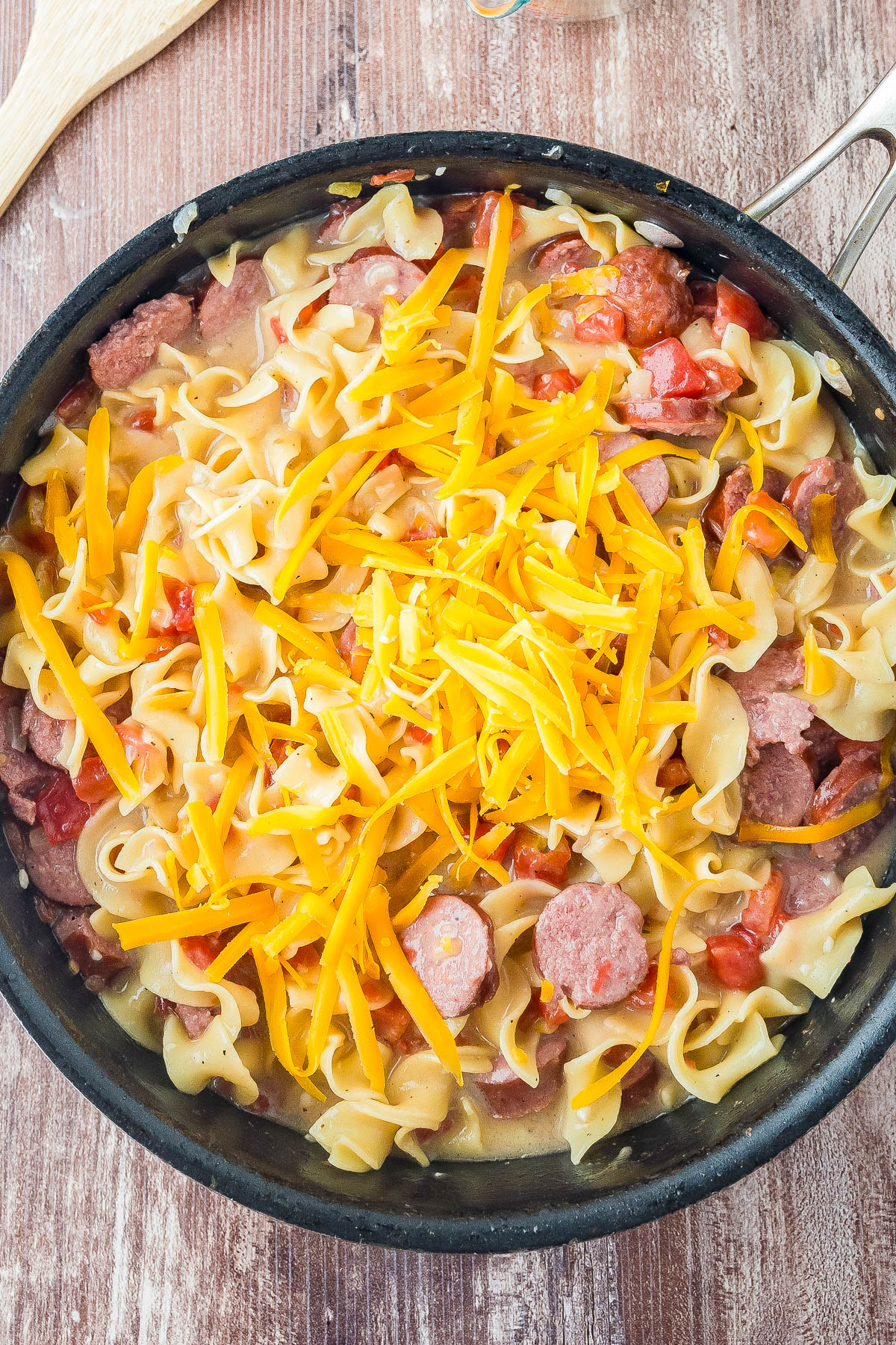 A skillet filled with cooked egg noodles, sliced sausage, diced tomatoes, and topped with shredded cheddar cheese.