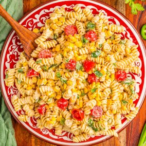 A plate of rotini pasta salad with cherry tomatoes, corn, green onions, cilantro, and a creamy dressing, surrounded by lime wedges, jalapeños, tomatoes, and spices on a wooden table.
