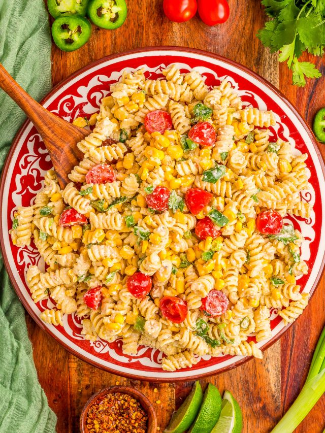 A plate of rotini pasta salad with cherry tomatoes, corn, green onions, cilantro, and a creamy dressing, surrounded by lime wedges, jalapeños, tomatoes, and spices on a wooden table.