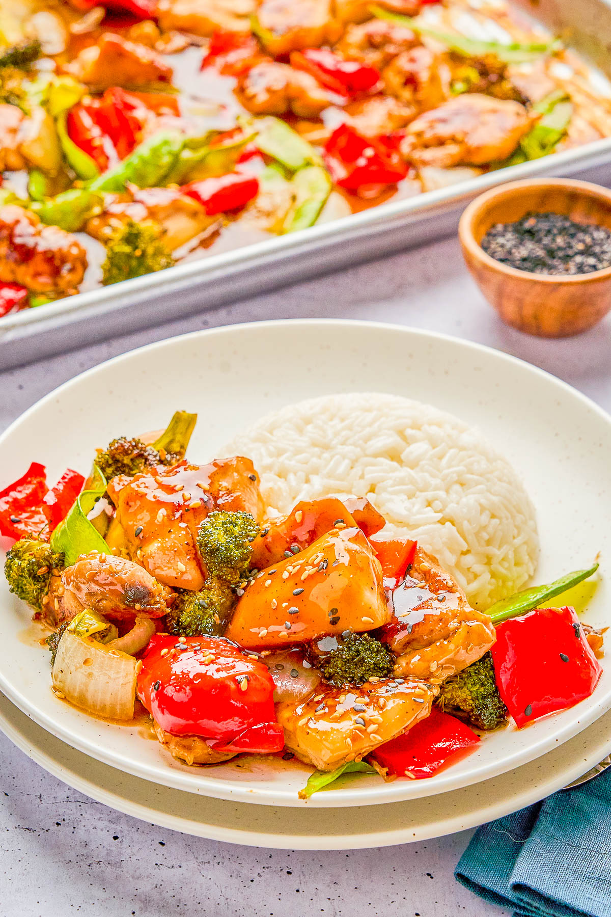 A plate of teriyaki chicken with red bell peppers, broccoli, onions, and a serving of white rice, garnished with sesame seeds. A tray with more of the same dish is in the background.