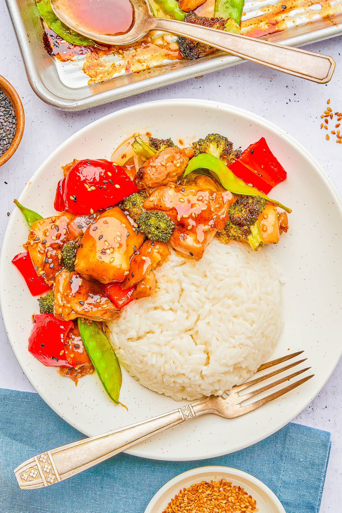 Plate of stir-fried chicken with broccoli, red peppers, and snap peas in sauce, served with rice. A fork is on the plate, and a spoon rests on a sauce-coated baking sheet.