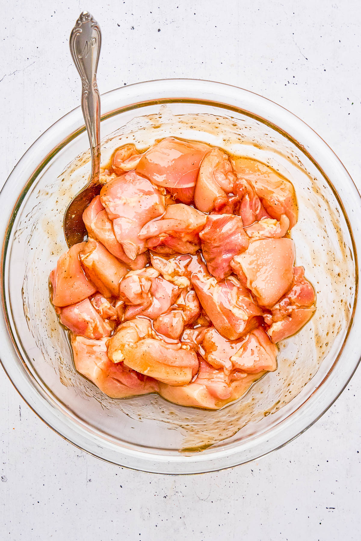 Raw marinated chicken pieces in a clear glass bowl with a metal spoon, on a white speckled surface.