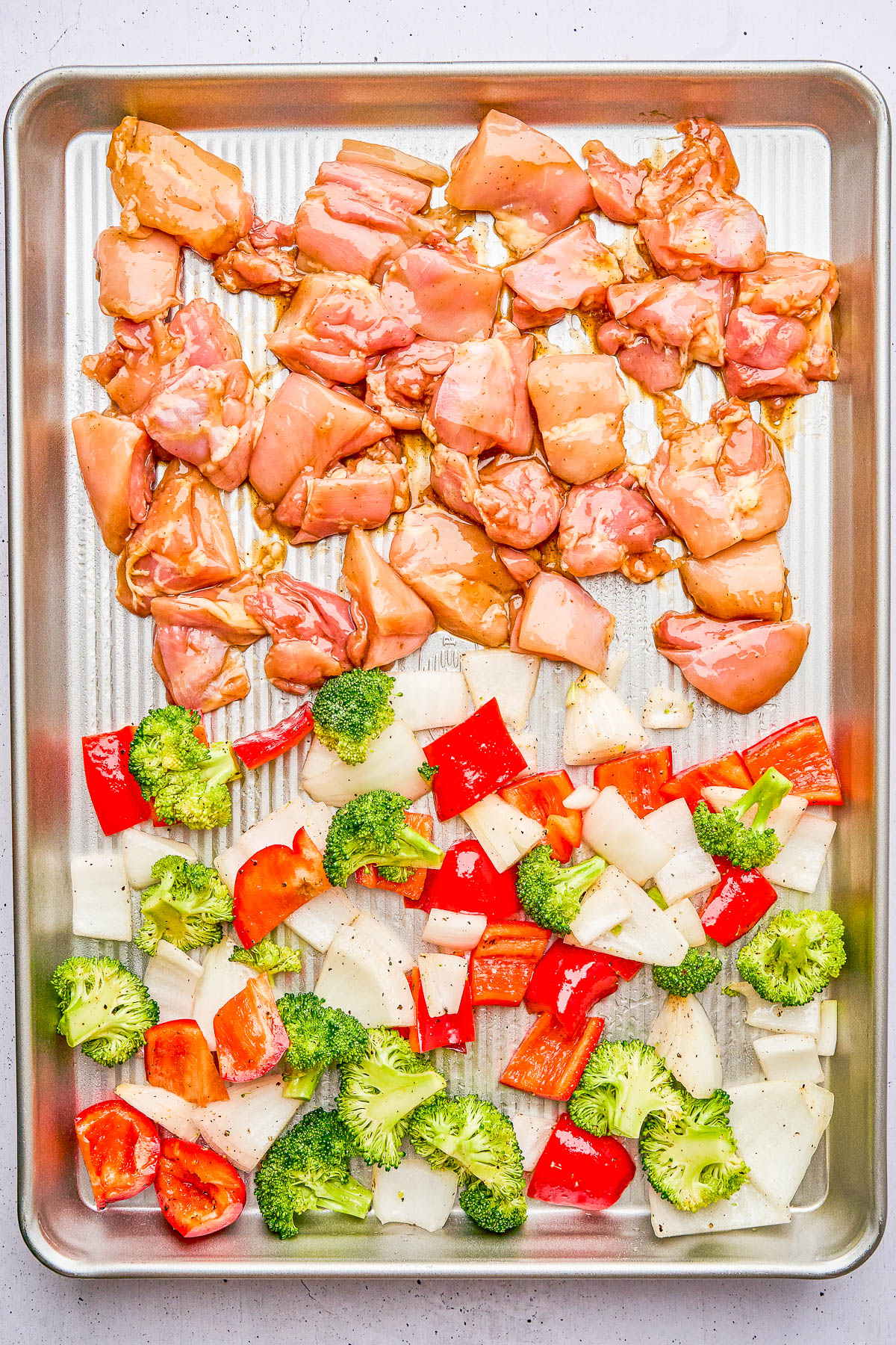 Sheet pan with raw diced chicken at the top and chopped vegetables, including broccoli, red peppers, and onions, at the bottom, ready for cooking.