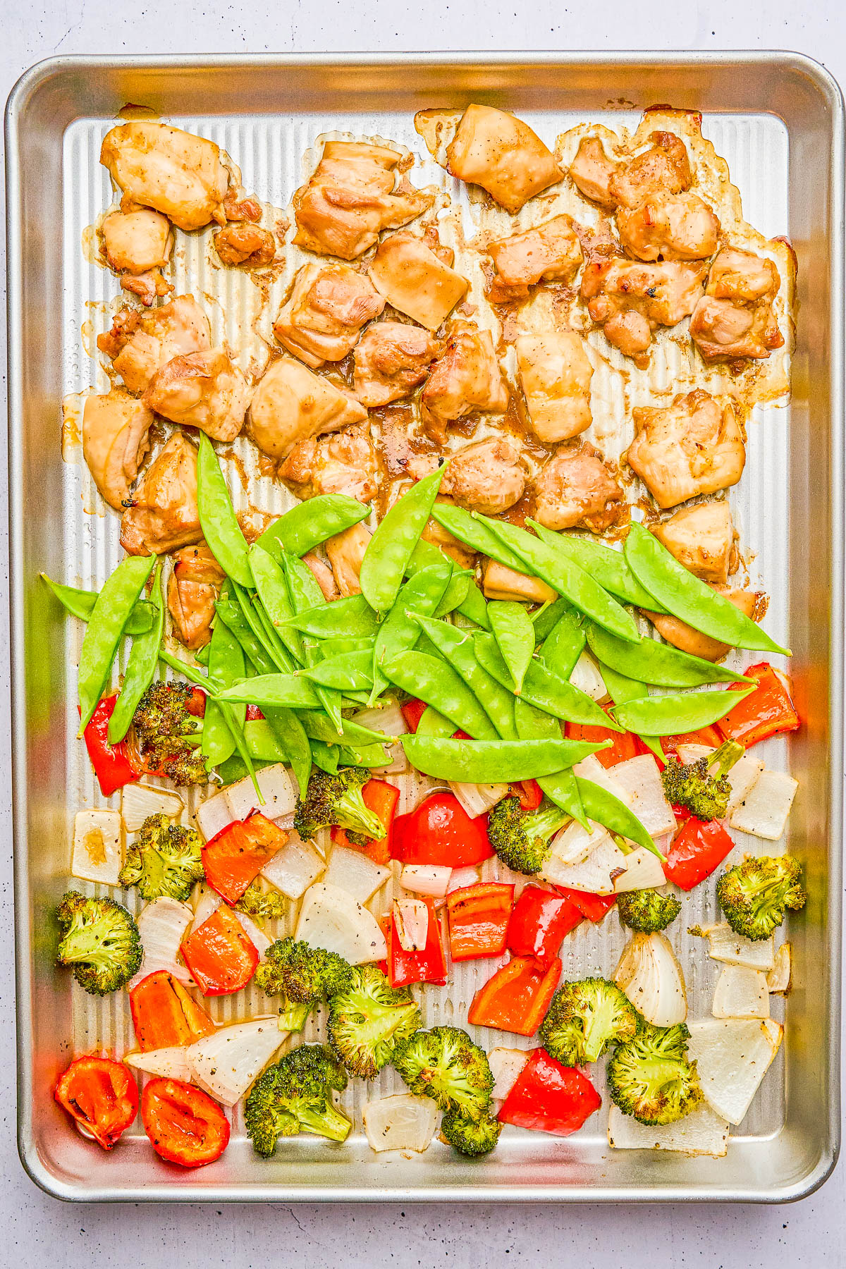 A baking sheet with cooked chicken pieces, snow peas, red bell peppers, broccoli, and onion, all arranged in separate sections.