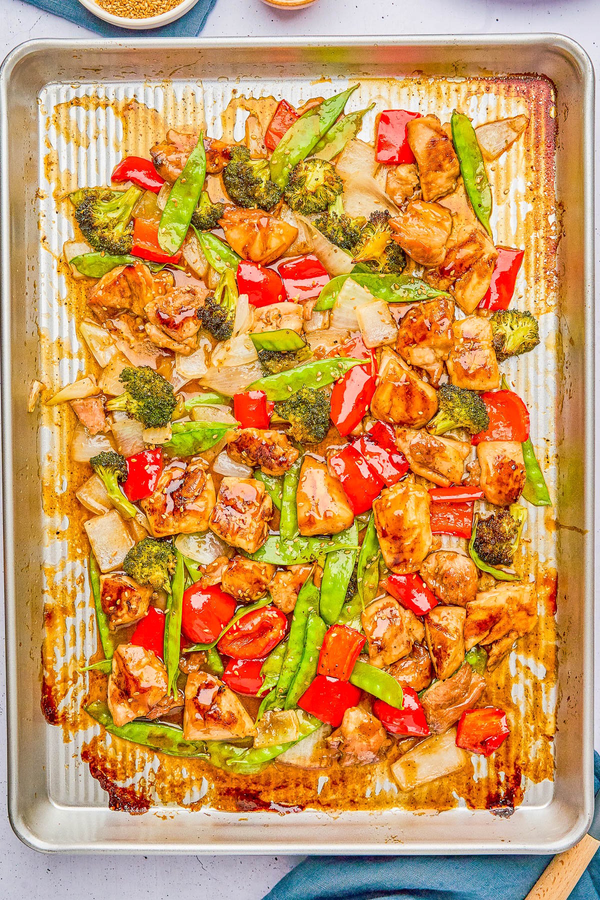 A baking sheet filled with cooked chicken pieces, red bell peppers, snap peas, broccoli, and onions, all coated in a sauce.