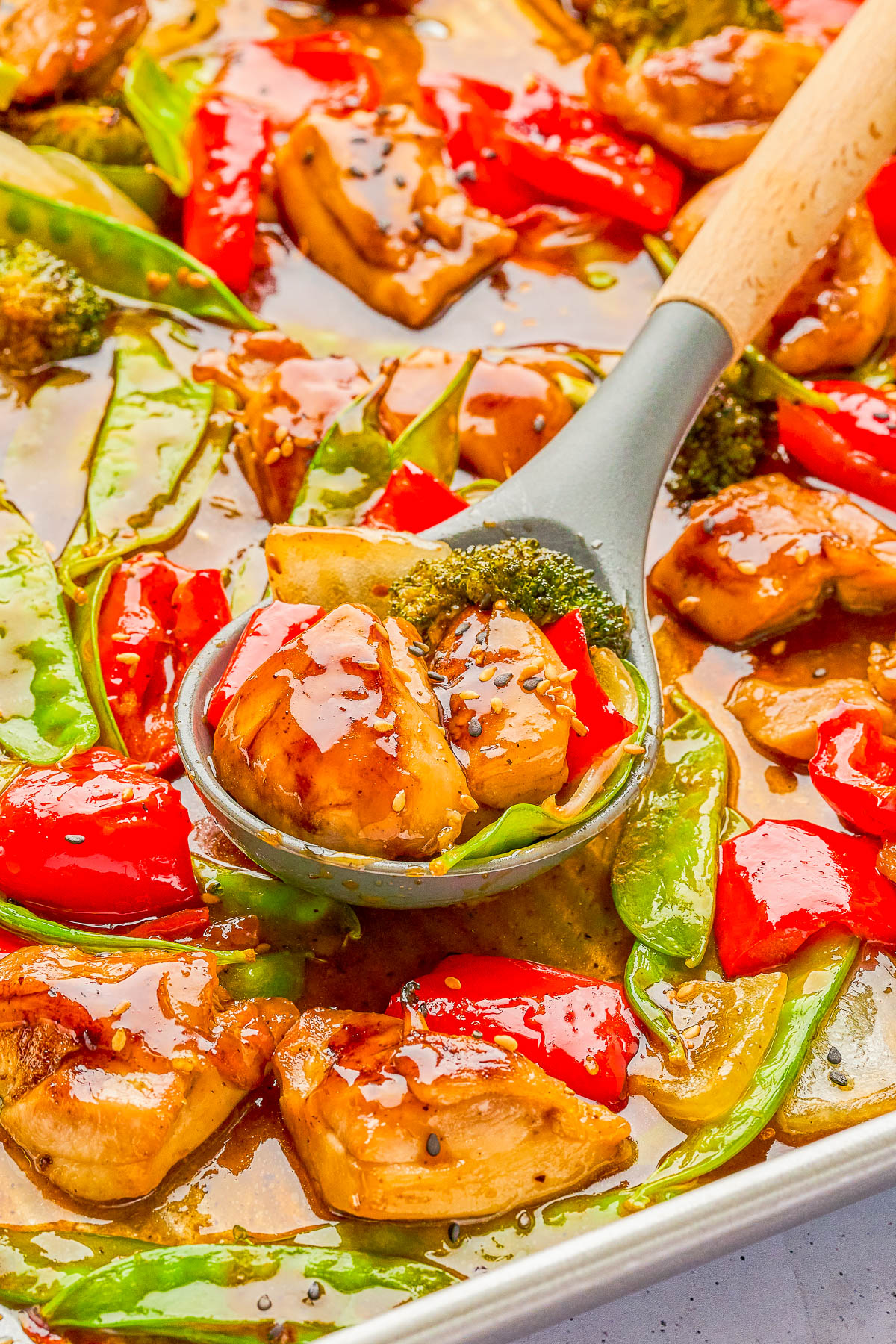 A spoon scoops glazed chicken, red bell peppers, snow peas, and broccoli from a baking sheet, with a glossy sauce coating the ingredients.