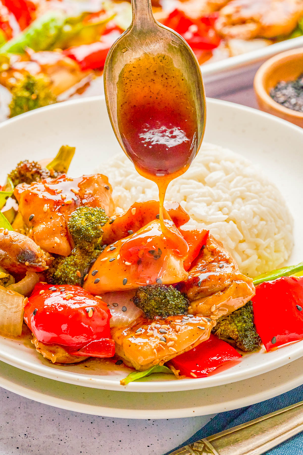 Spoon drizzling sauce onto a plate of chicken stir-fry with broccoli, red peppers, and white rice.