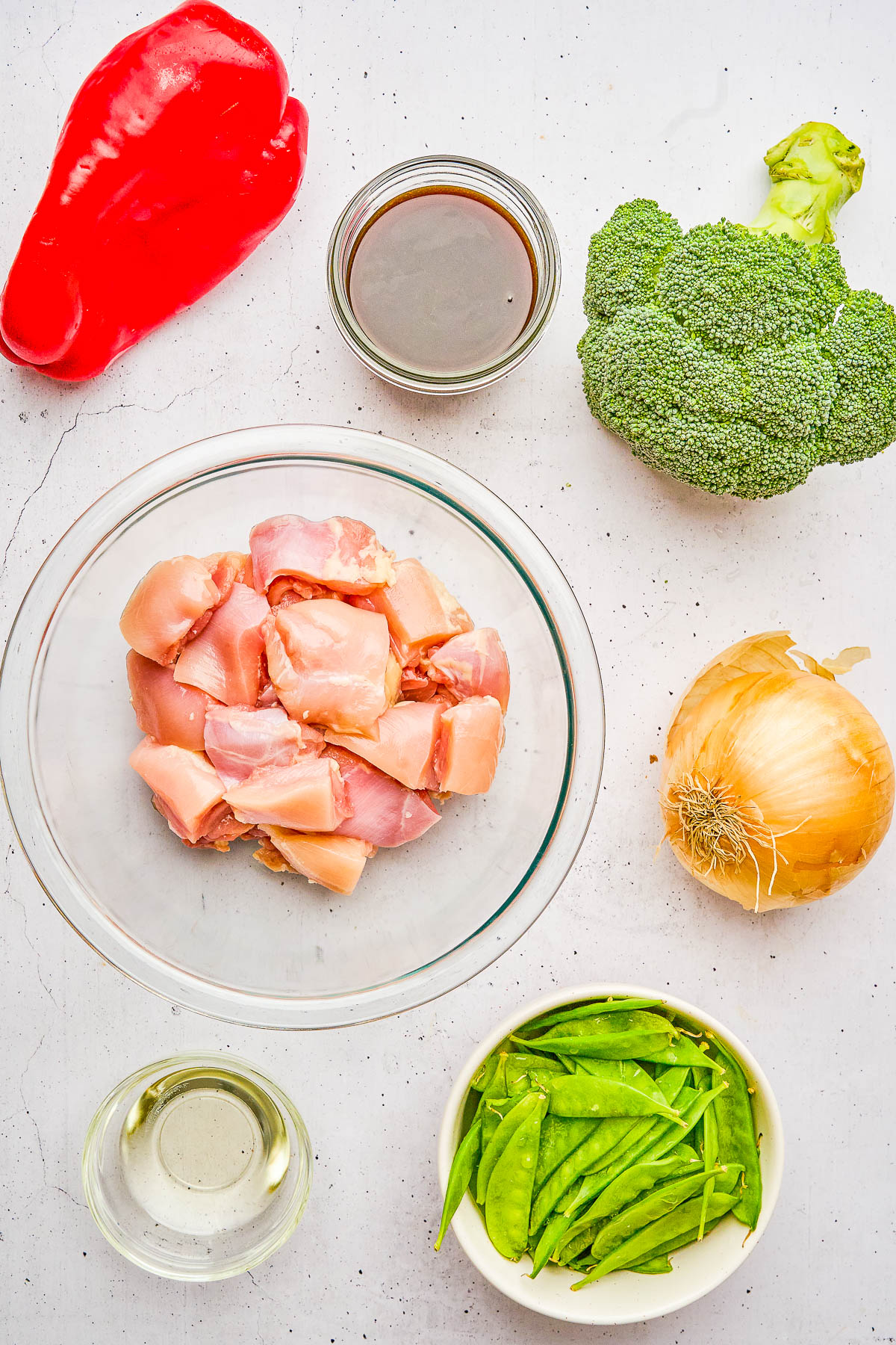 Raw chicken pieces, red bell pepper, broccoli, onion, snow peas, and sauce ingredients are arranged on a white surface in bowls and jars.