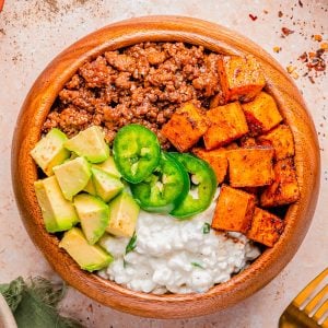A wooden bowl containing seasoned ground meat, roasted sweet potatoes, diced avocado, cottage cheese, and sliced jalapeño, arranged in sections.