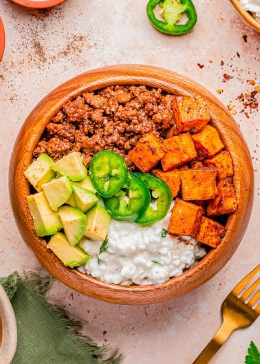 A wooden bowl containing seasoned ground meat, roasted sweet potatoes, diced avocado, cottage cheese, and sliced jalapeño, arranged in sections.