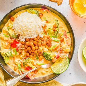 A plate of curry with broccoli, peppers, rice, crispy chickpeas, and sliced lime, surrounded by small bowls of herbs, rice, and a glass of iced citrus drink.