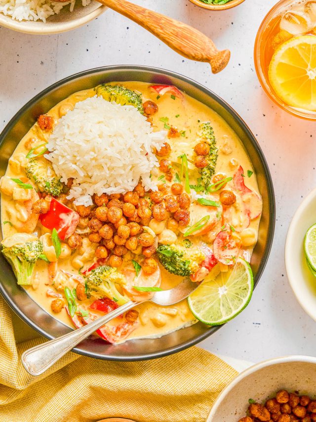 A plate of curry with broccoli, peppers, rice, crispy chickpeas, and sliced lime, surrounded by small bowls of herbs, rice, and a glass of iced citrus drink.