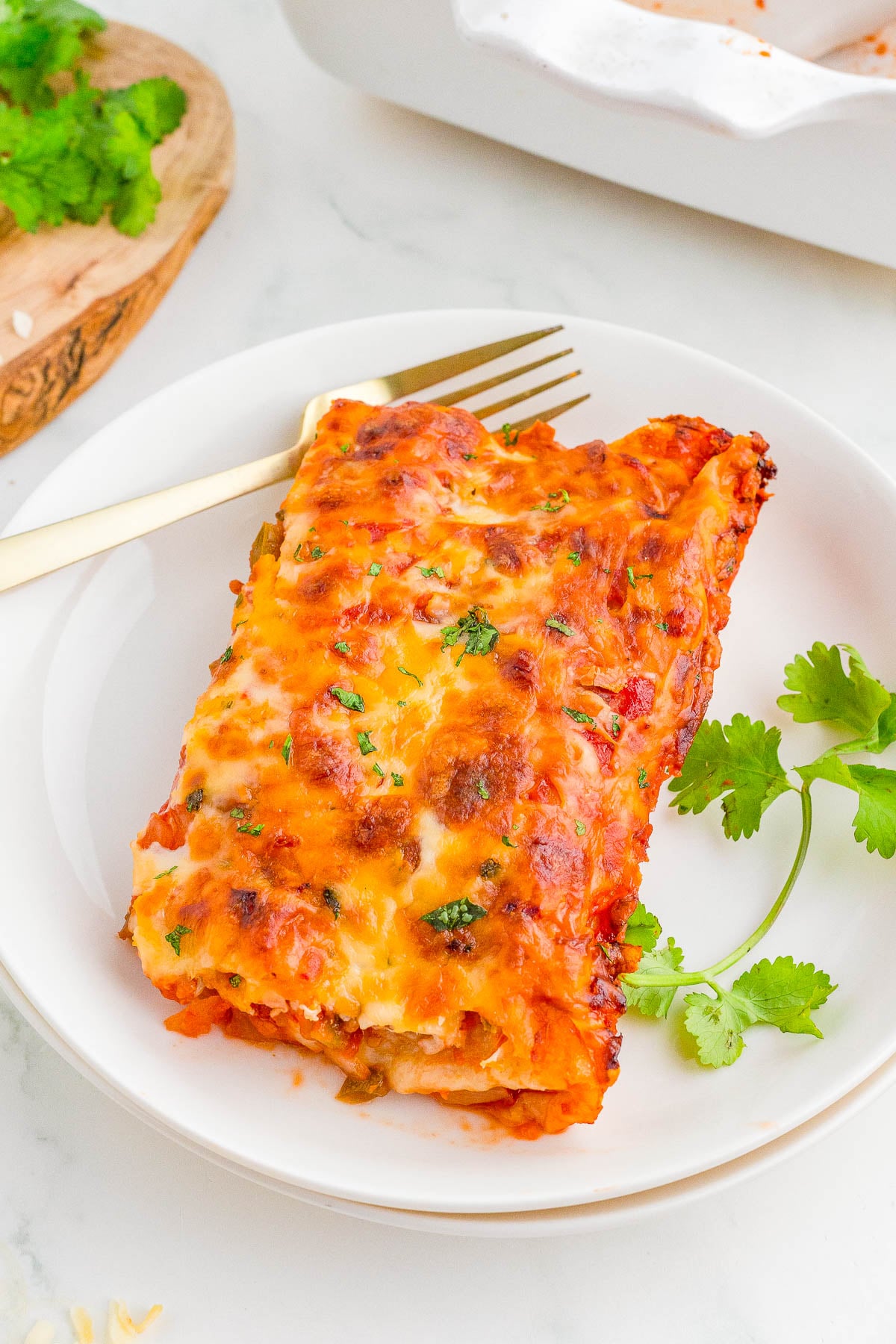 A serving of cheesy baked enchiladas on a white plate, garnished with fresh cilantro, with a gold fork beside the food.