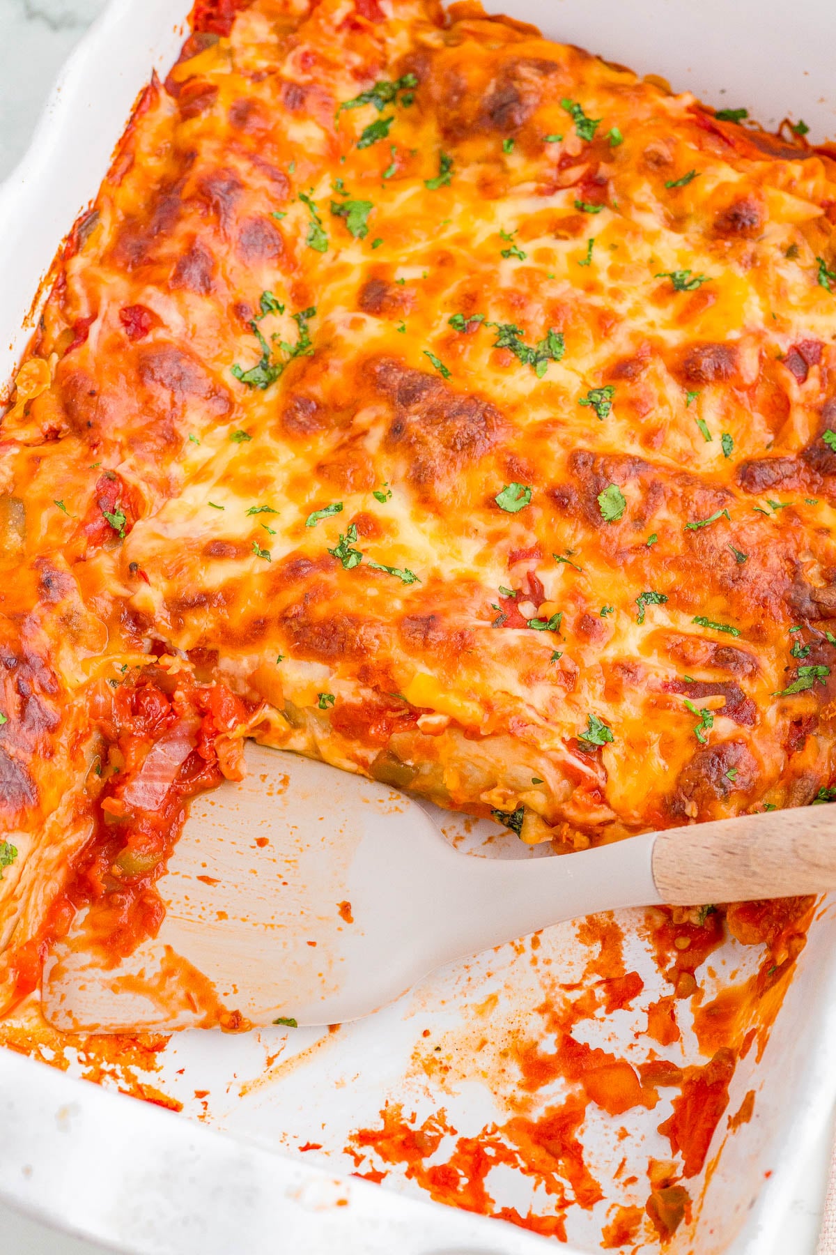 A partially served baked lasagna in a white dish with a wooden spatula, garnished with chopped herbs.