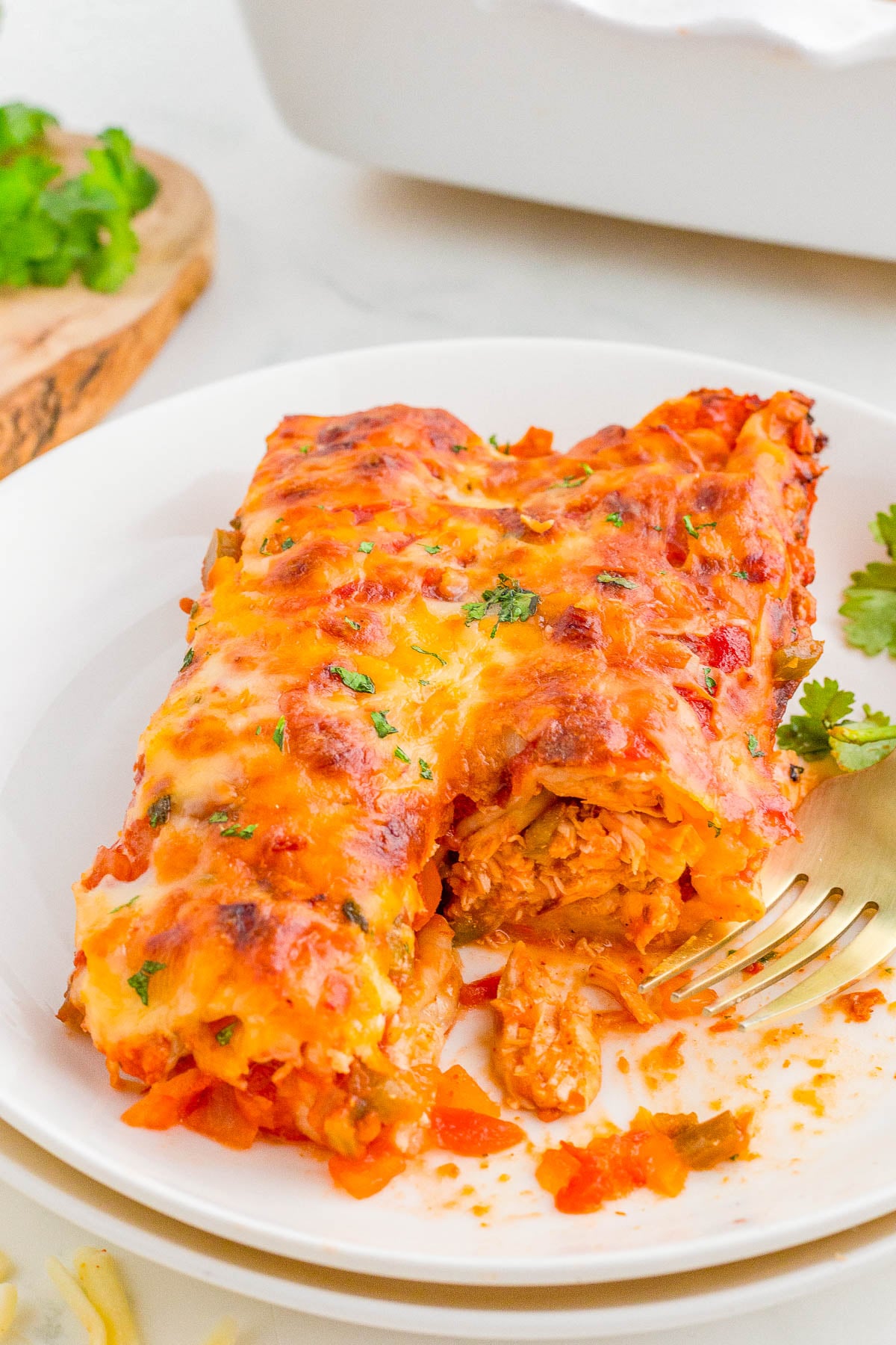 A serving of cheesy baked enchiladas filled with shredded chicken and vegetables is on a white plate with a fork and garnish.