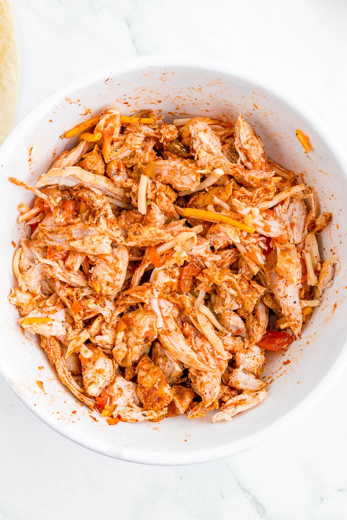 A white bowl filled with shredded chicken mixed with spices, shredded cheese, and small pieces of vegetables.