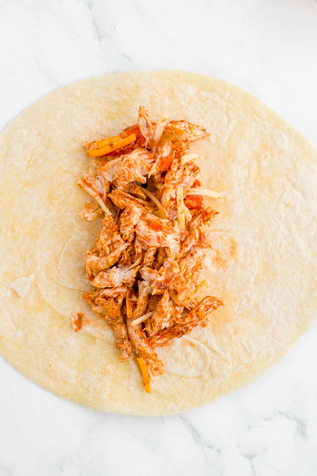 Shredded chicken with spices and onions placed in the center of a flour tortilla on a white surface.