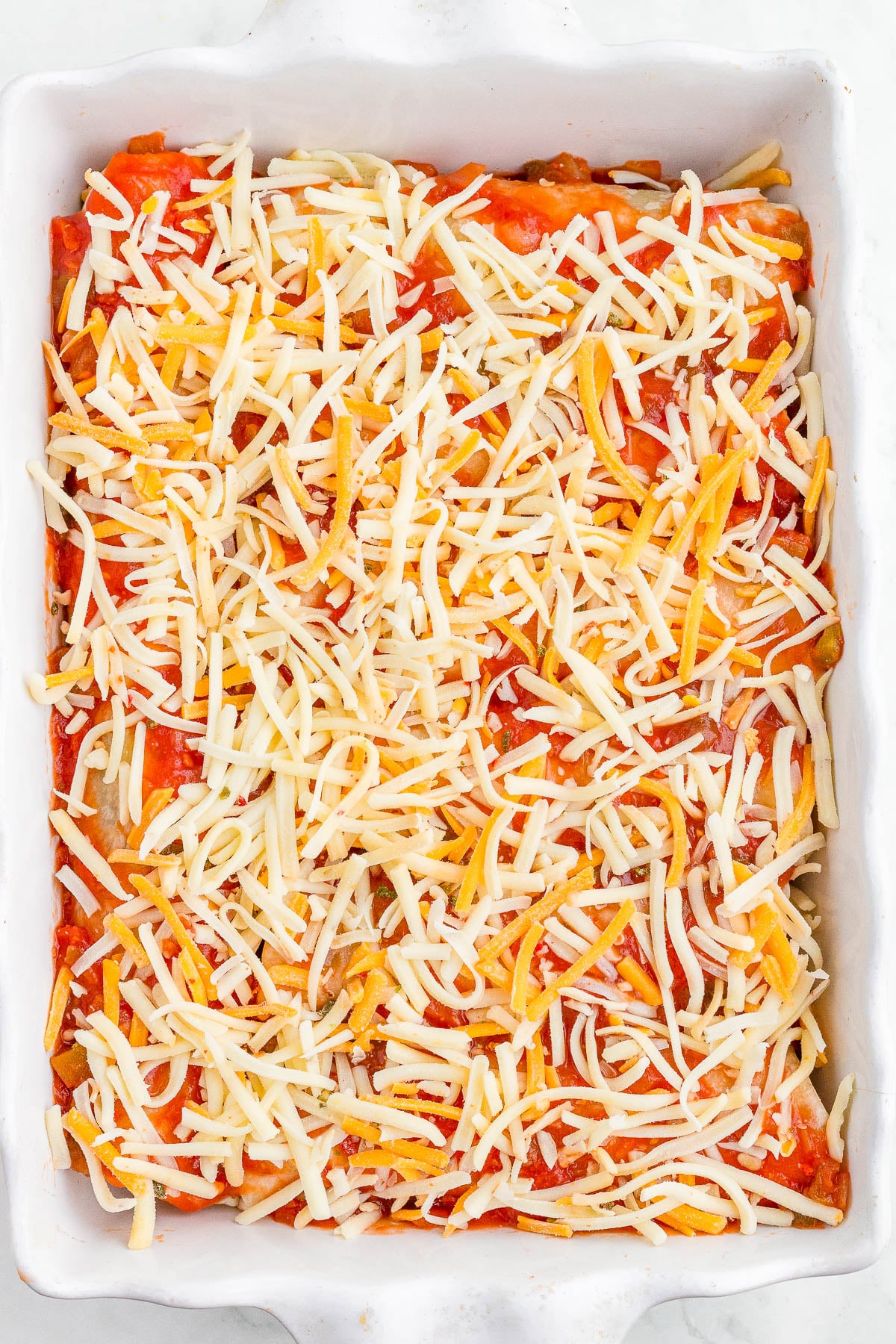 A white baking dish filled with an uncooked casserole topped with shredded cheese and diced tomatoes.
