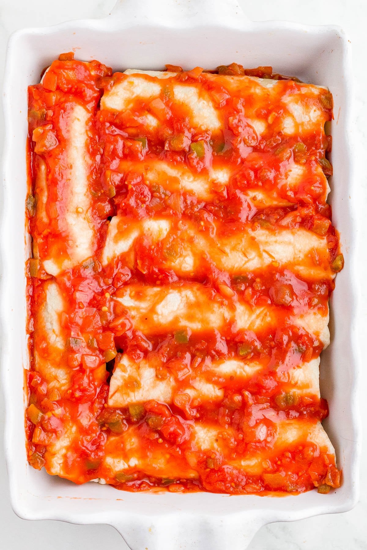 A white baking dish filled with rolled enchiladas topped with chunky red sauce and diced vegetables, ready to be baked.