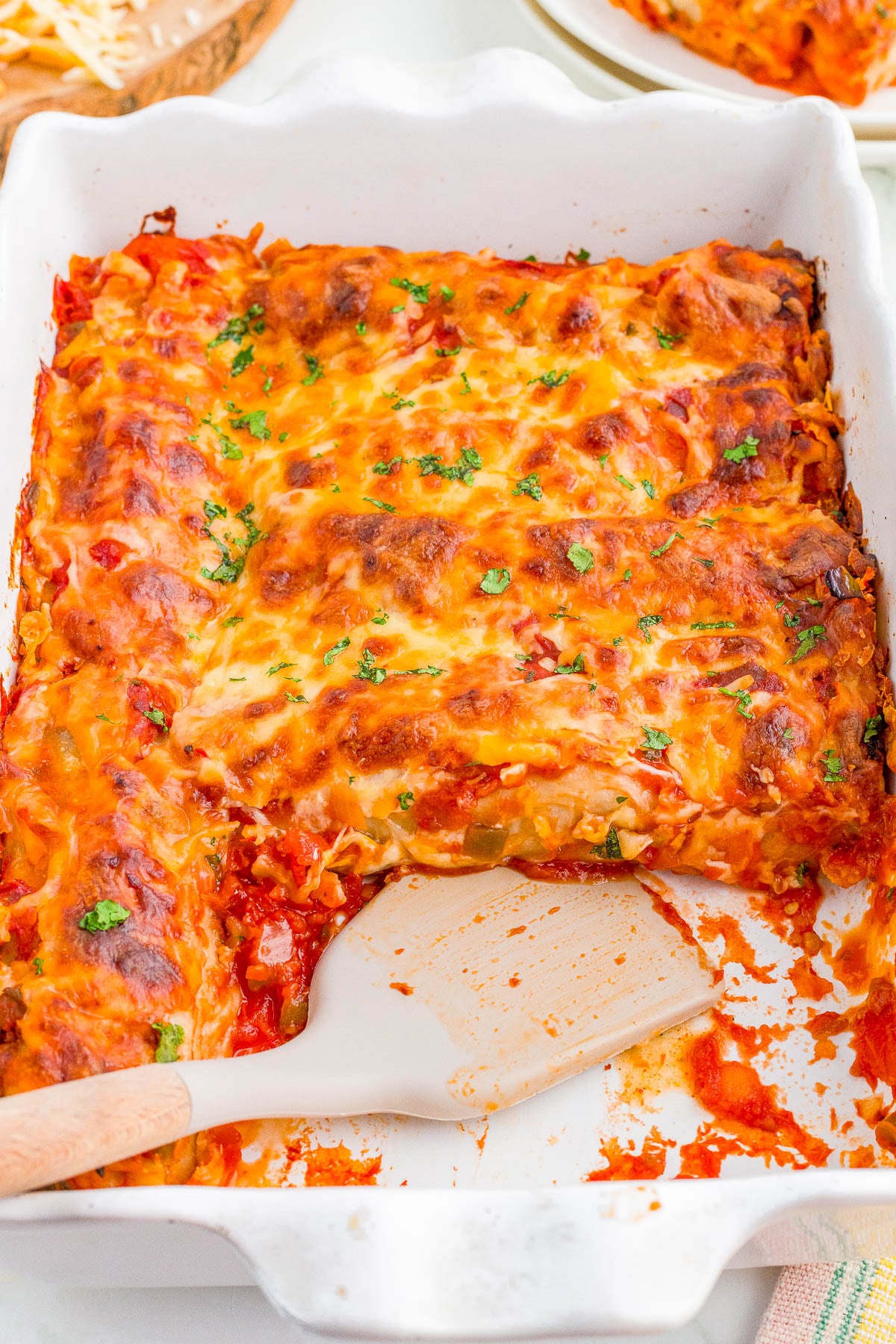 A baked casserole dish of cheesy enchiladas with a serving removed, revealing the interior, and a spatula resting in the dish.