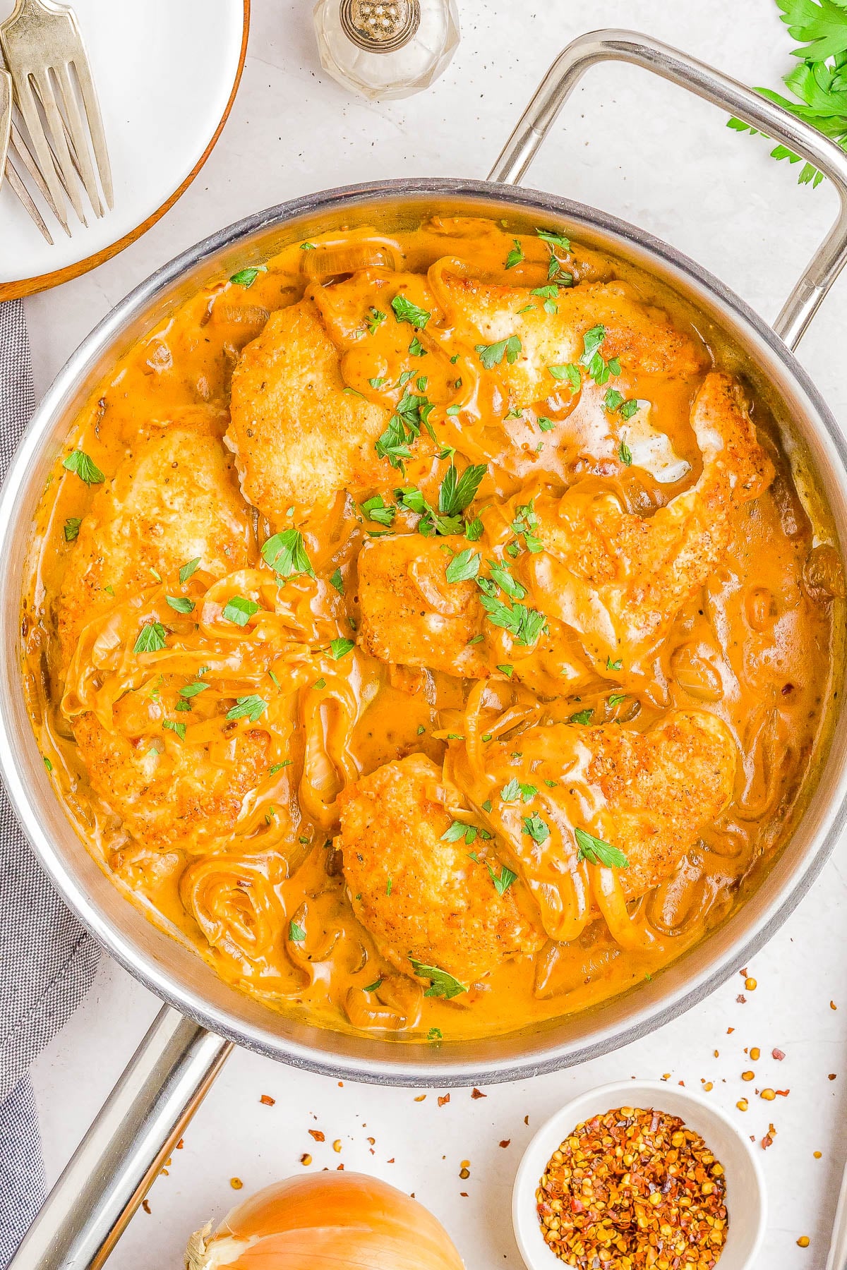 A skillet filled with breaded chicken breasts in a creamy orange sauce with sliced onions, garnished with chopped parsley.