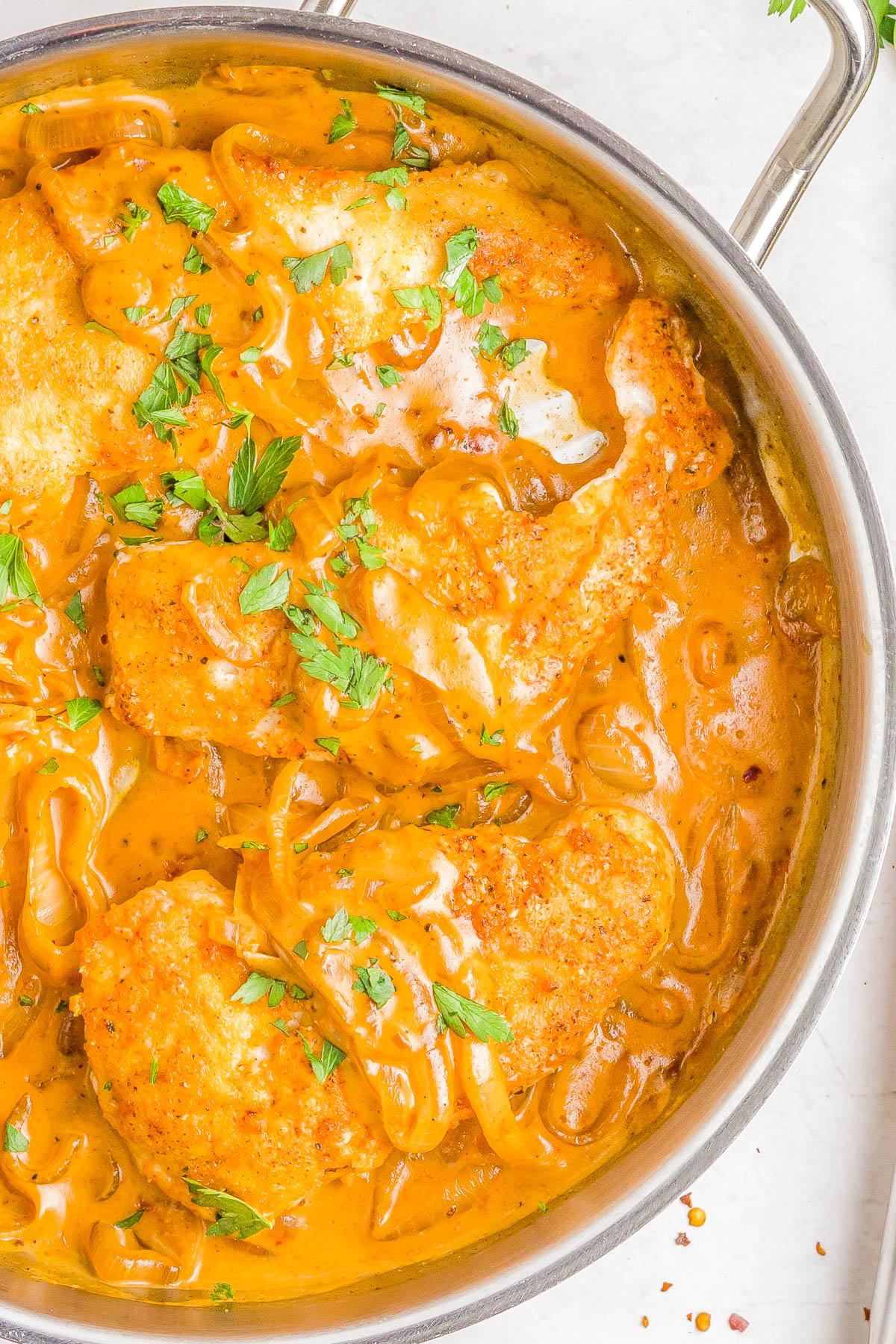 Pan-seared chicken breasts in a creamy orange-brown sauce with sliced onions, garnished with chopped parsley in a stainless steel pan.