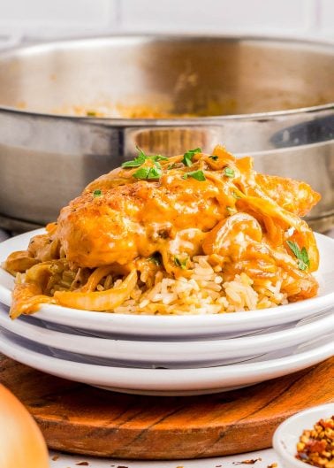 A serving of creamy chicken with sauce and herbs over rice on a white plate, with a pan of the same dish in the background.