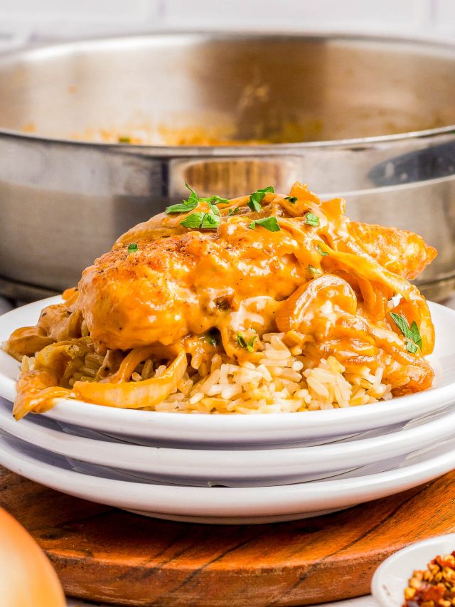 A serving of creamy chicken with sauce and herbs over rice on a white plate, with a pan of the same dish in the background.