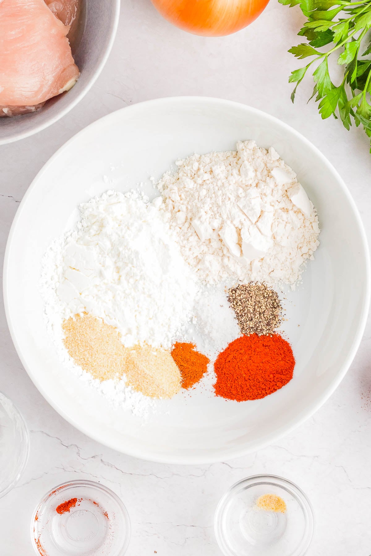A white bowl containing flour, cornstarch, paprika, black pepper, garlic powder, and onion powder, with parsley, onion, and raw chicken nearby.
