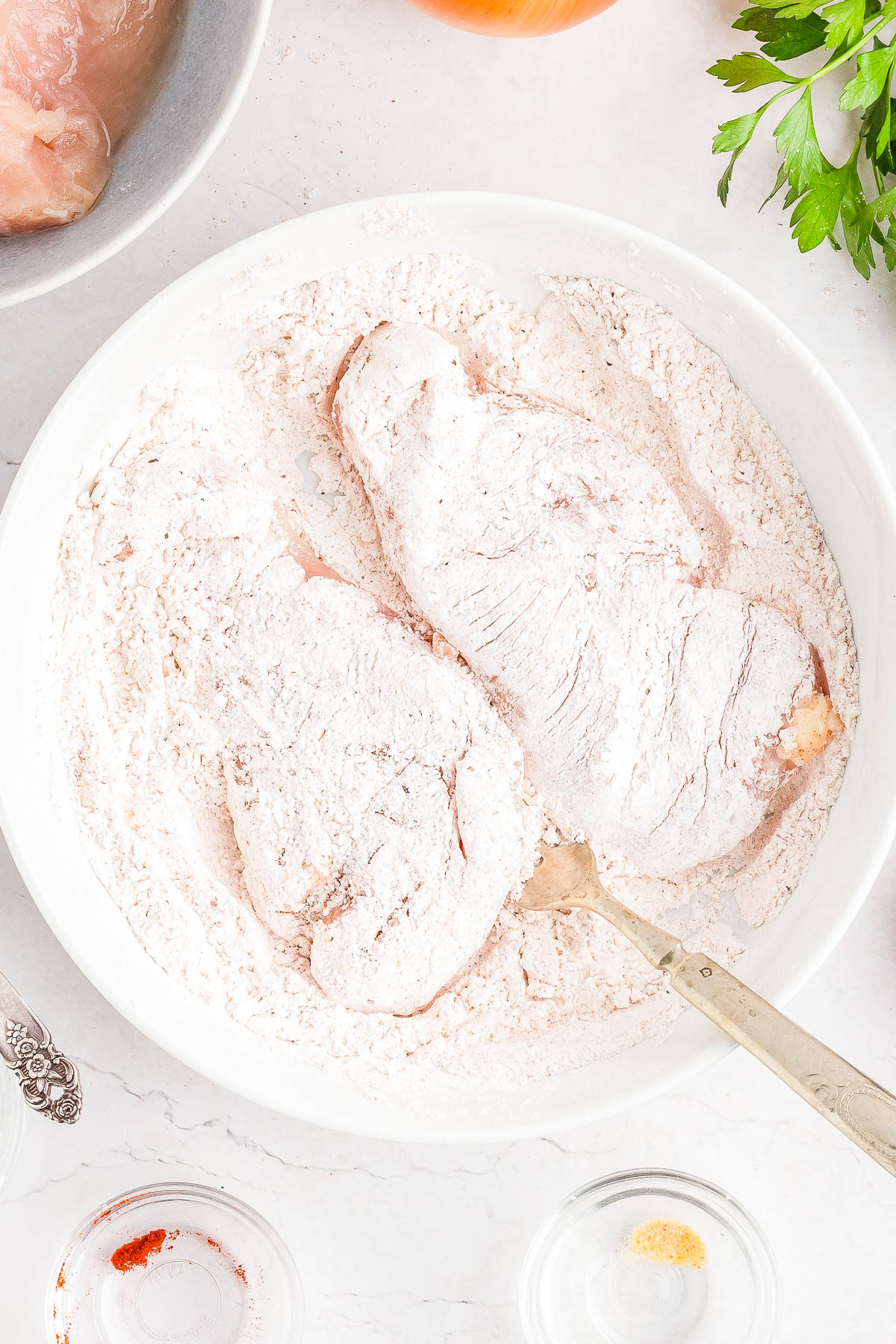 Two raw chicken breasts coated in flour sit in a white bowl with a fork, surrounded by ingredients on a white countertop.