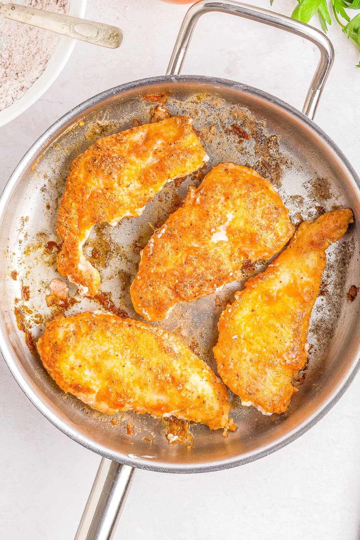 Four breaded chicken breasts cooking in a stainless steel frying pan on a white surface.