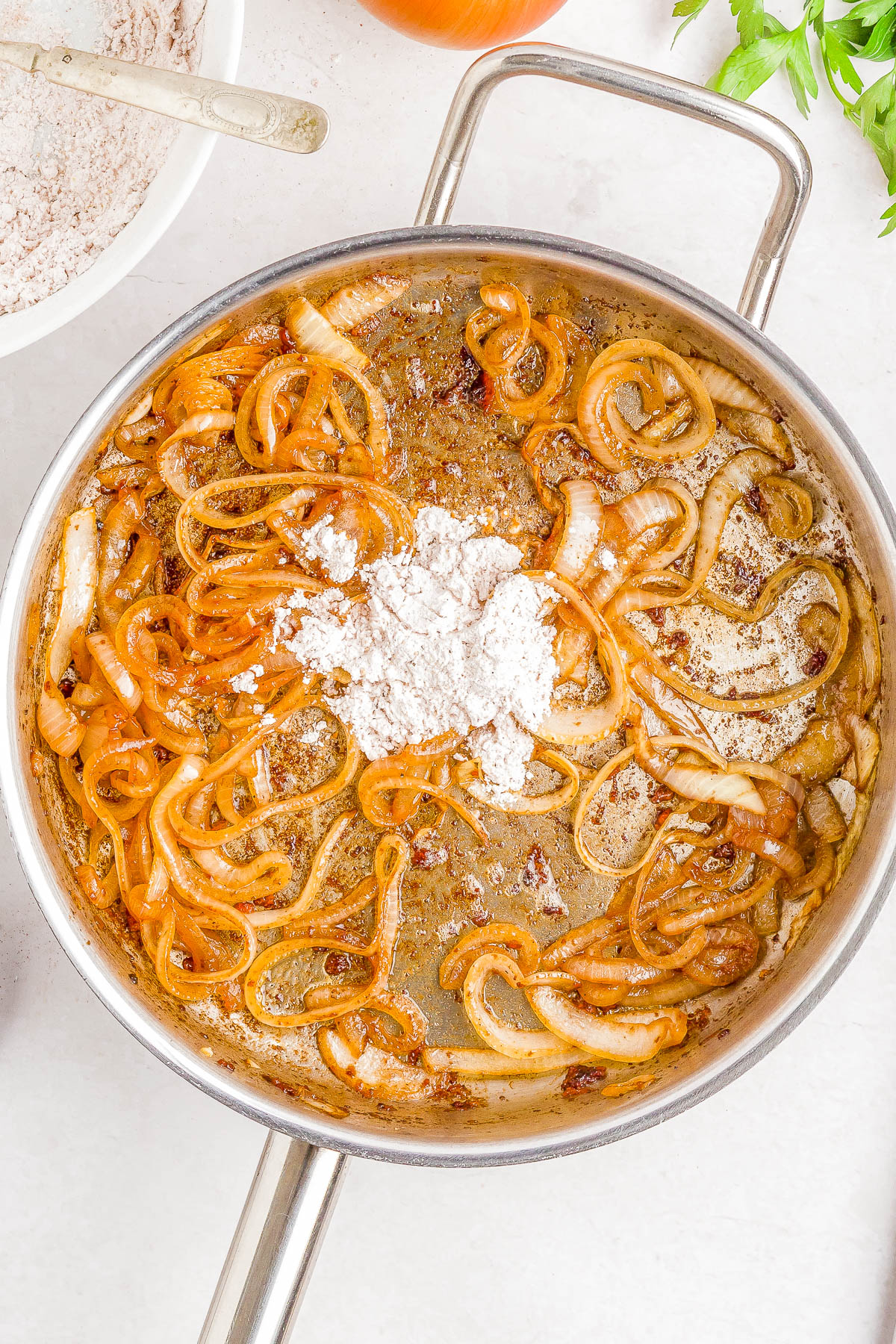 Sliced onions cooking in a stainless steel pan with a mound of flour added on top, surrounded by other kitchen ingredients.