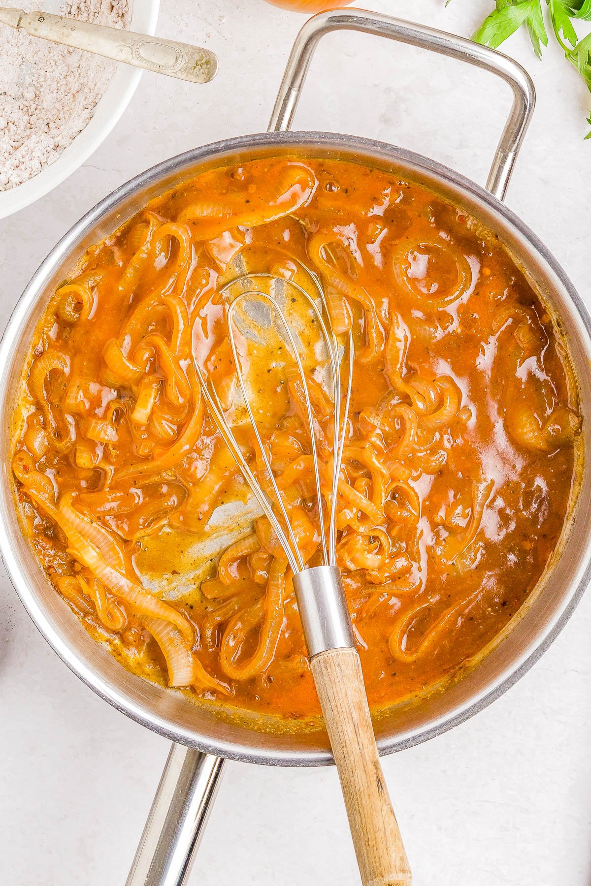 A stainless steel pan filled with onion gravy being stirred with a metal whisk; sliced onions are visible in the thick brown sauce.