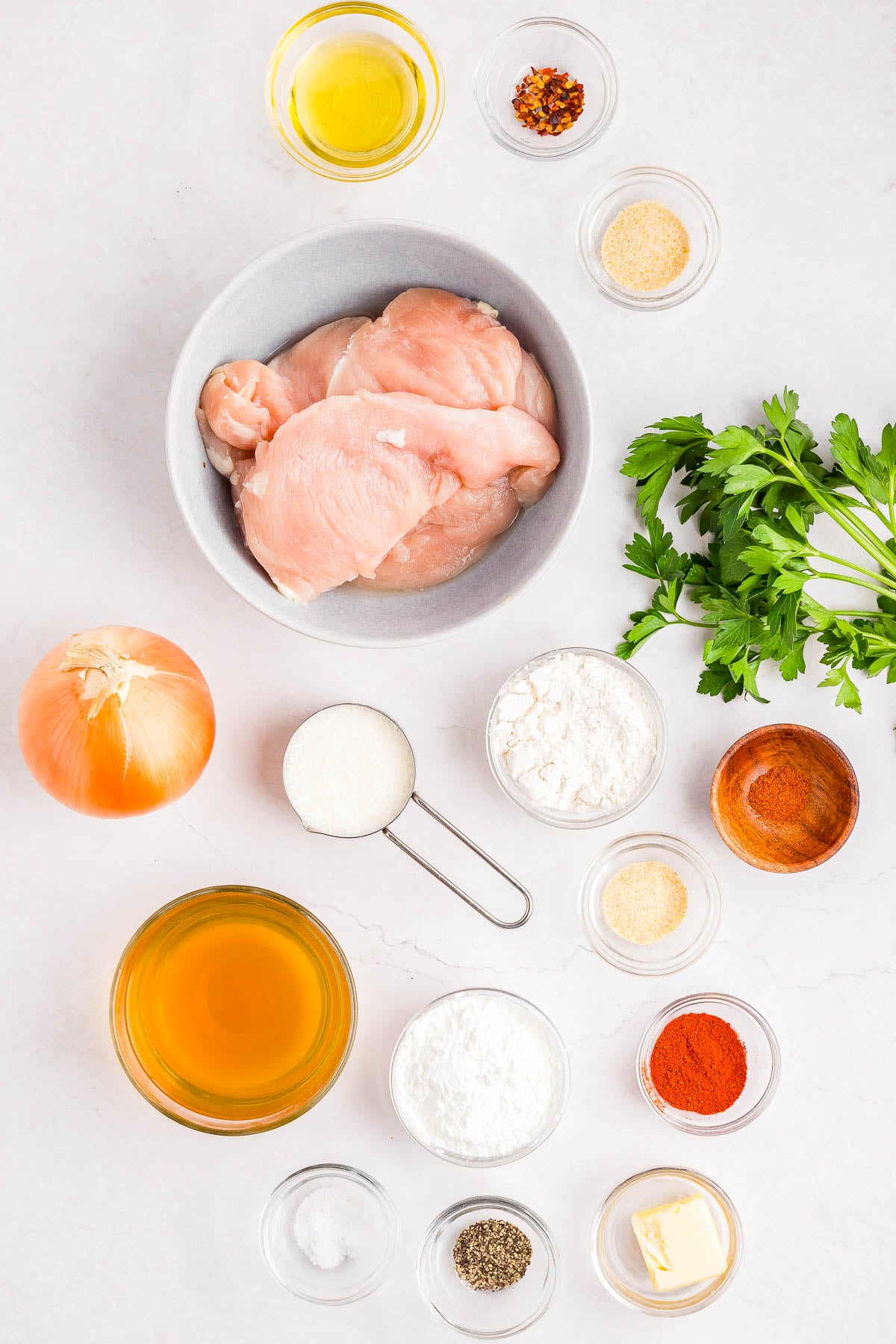Various raw ingredients arranged on a white surface, including chicken breast, onion, parsley, flour, cornstarch, spices, oil, broth, butter, and measuring cups.