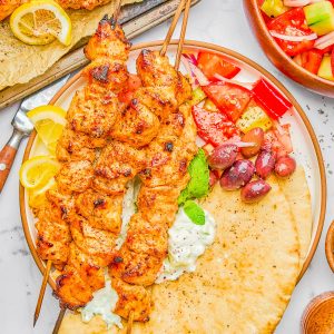 Grilled chicken skewers on pita bread with tzatziki, lemon wedges, olives, and a tomato-cucumber salad, served on a white plate.