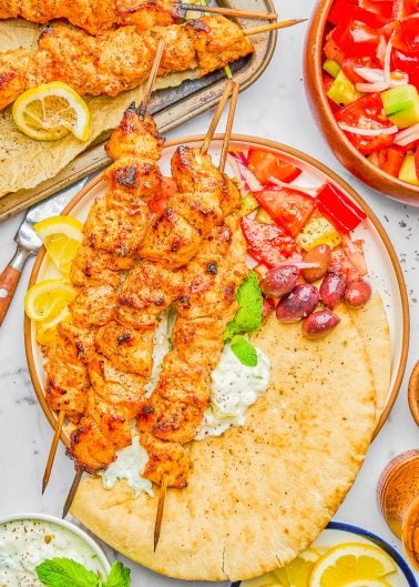 Grilled chicken skewers on pita bread with tzatziki, lemon wedges, olives, and a tomato-cucumber salad, served on a white plate.