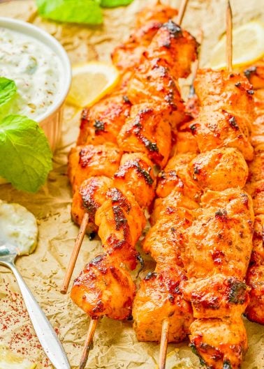 Grilled chicken skewers with a reddish marinade are arranged on parchment paper, accompanied by a bowl of yogurt sauce, mint leaves, and lemon slices.