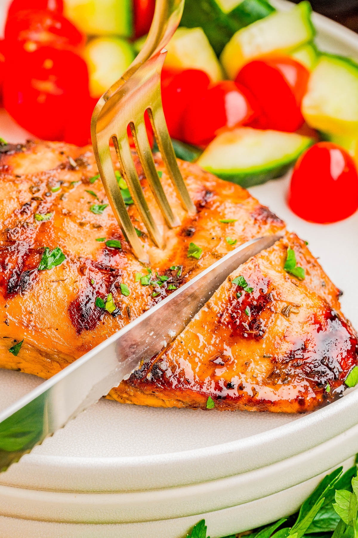 A fork and knife cut into a grilled chicken breast on a plate, served with sliced cucumbers and cherry tomatoes.