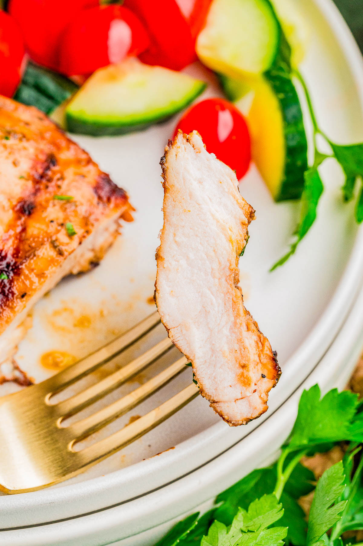 A close-up of a fork holding a slice of grilled chicken, with sliced cucumbers, tomatoes, and herbs on a plate in the background.