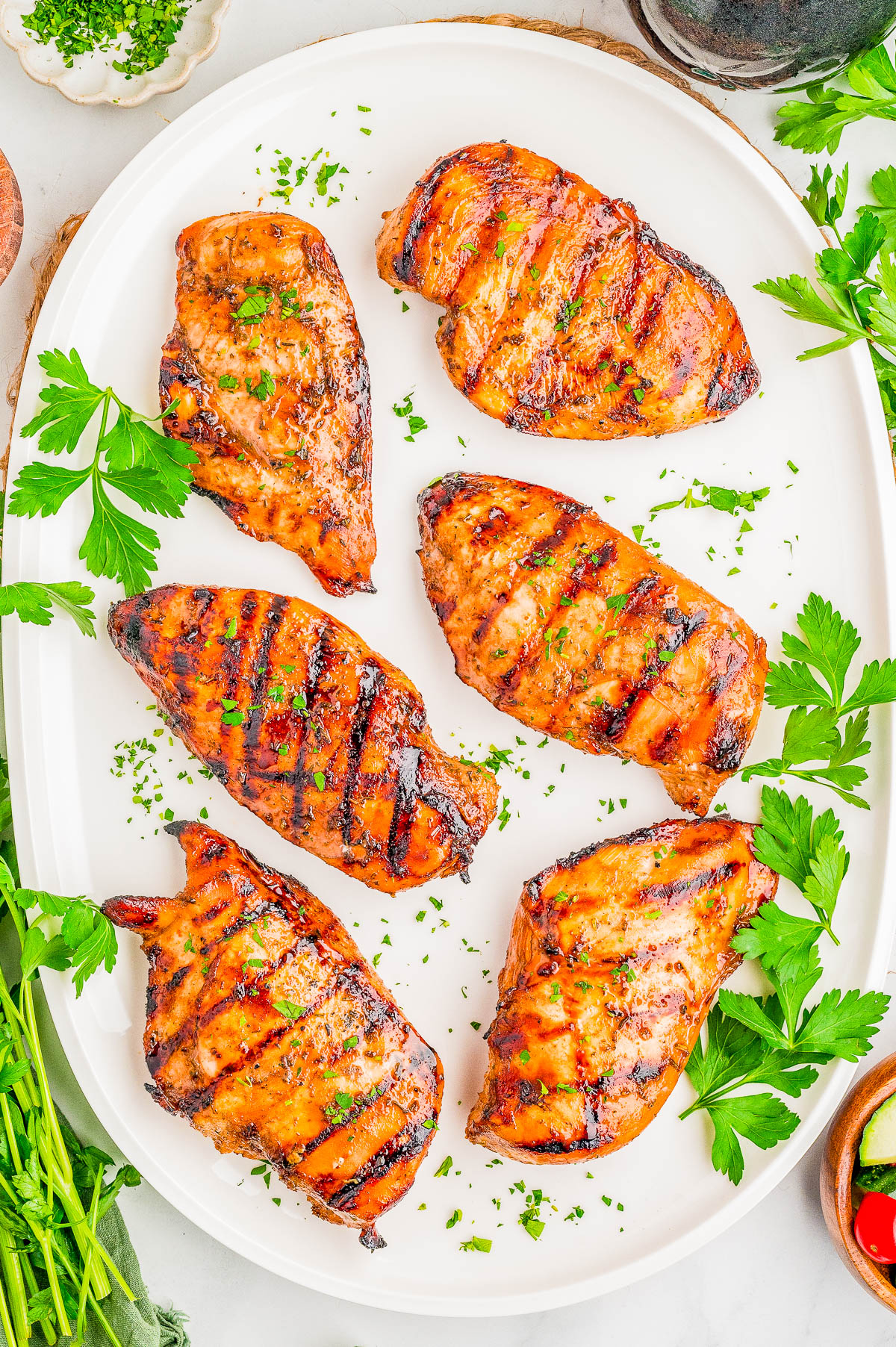 Six grilled chicken breasts with grill marks, garnished with chopped parsley, arranged on a white oval platter with parsley sprigs around the edges.