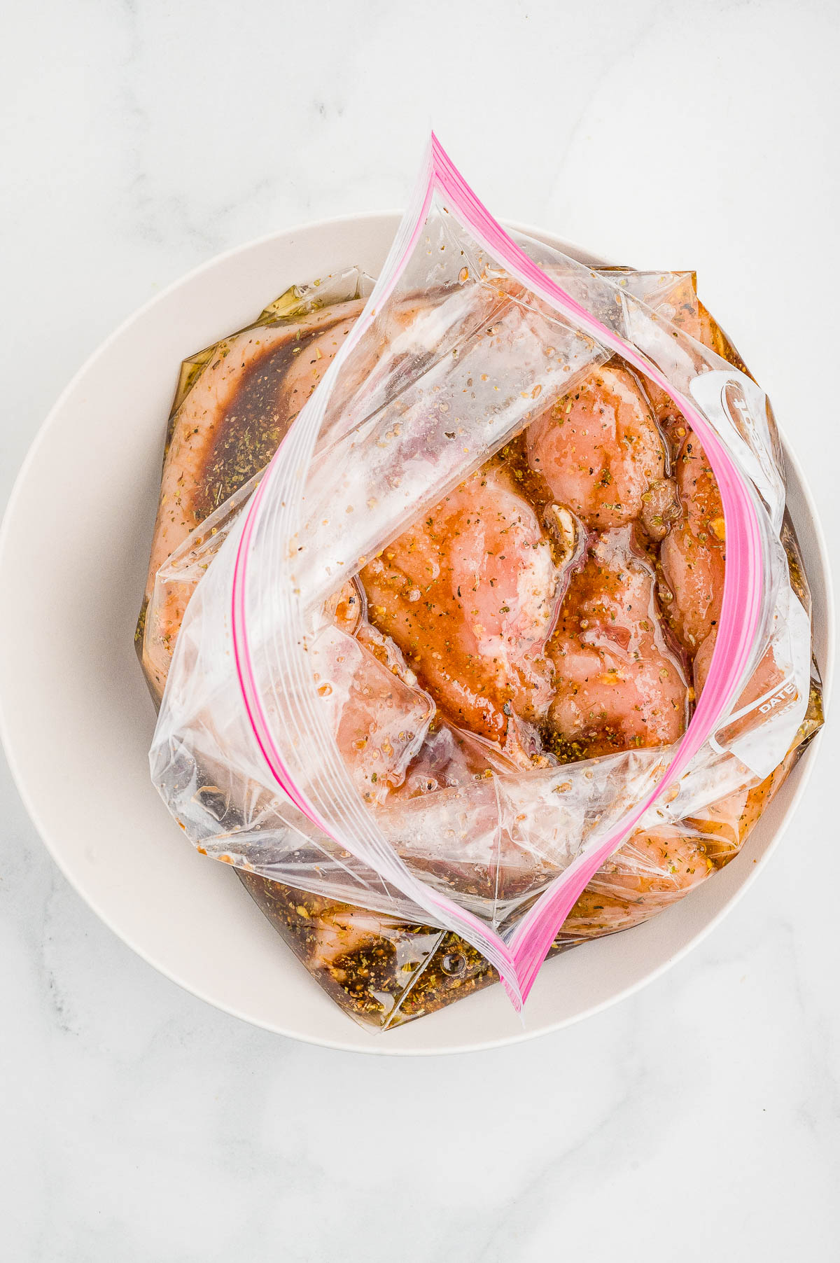Raw chicken pieces marinating in a clear plastic zip-top bag with dark marinade, placed in a white bowl on a white surface.