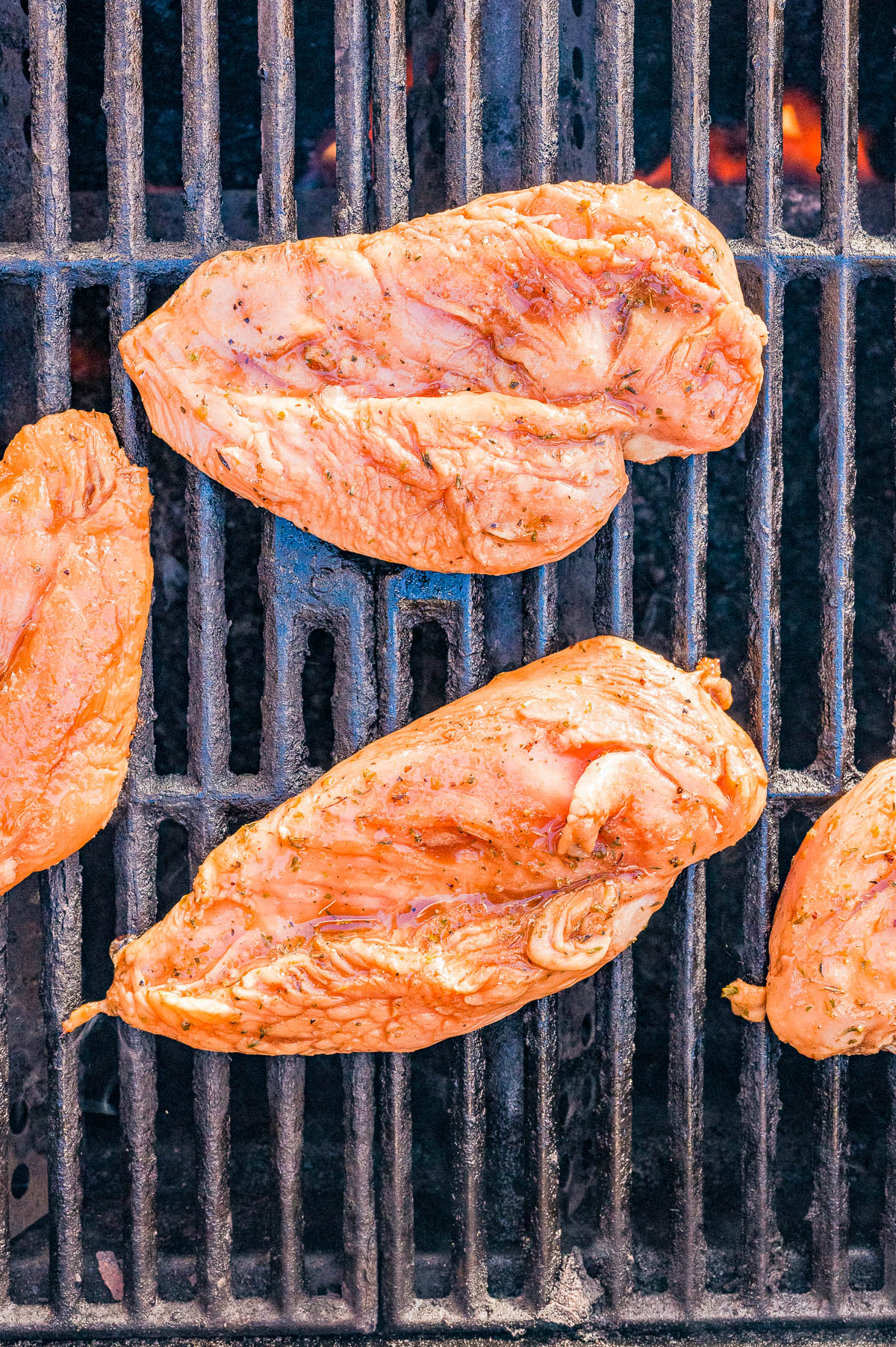 Raw, seasoned chicken breasts cooking on a grill with visible grill grates and flames underneath.