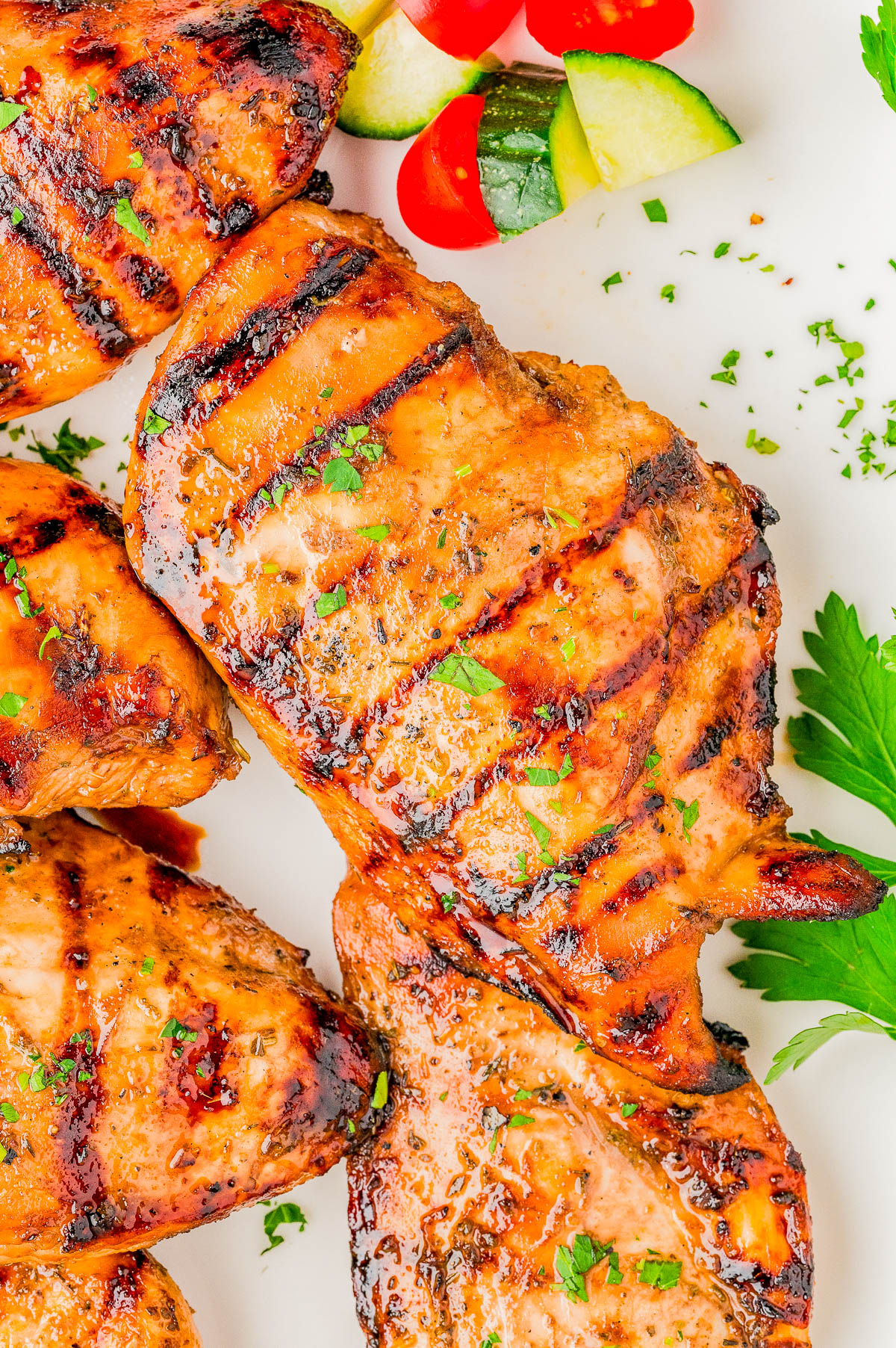 Grilled chicken breasts with visible char marks, garnished with chopped herbs, served alongside sliced cucumber and cherry tomatoes on a white plate.