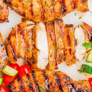Grilled chicken breasts, some sliced, garnished with herbs and served with cherry tomatoes and cucumber pieces on a white surface.