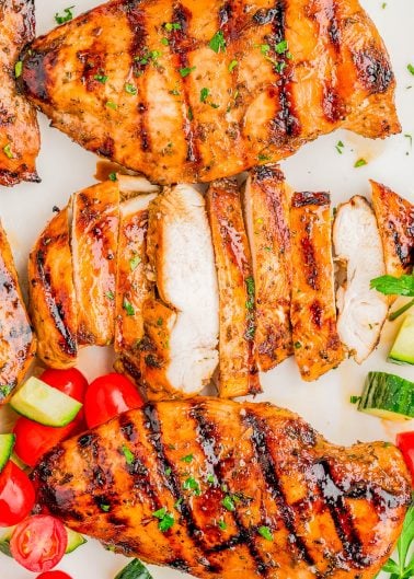 Grilled chicken breasts, some sliced, garnished with herbs and served with cherry tomatoes and cucumber pieces on a white surface.
