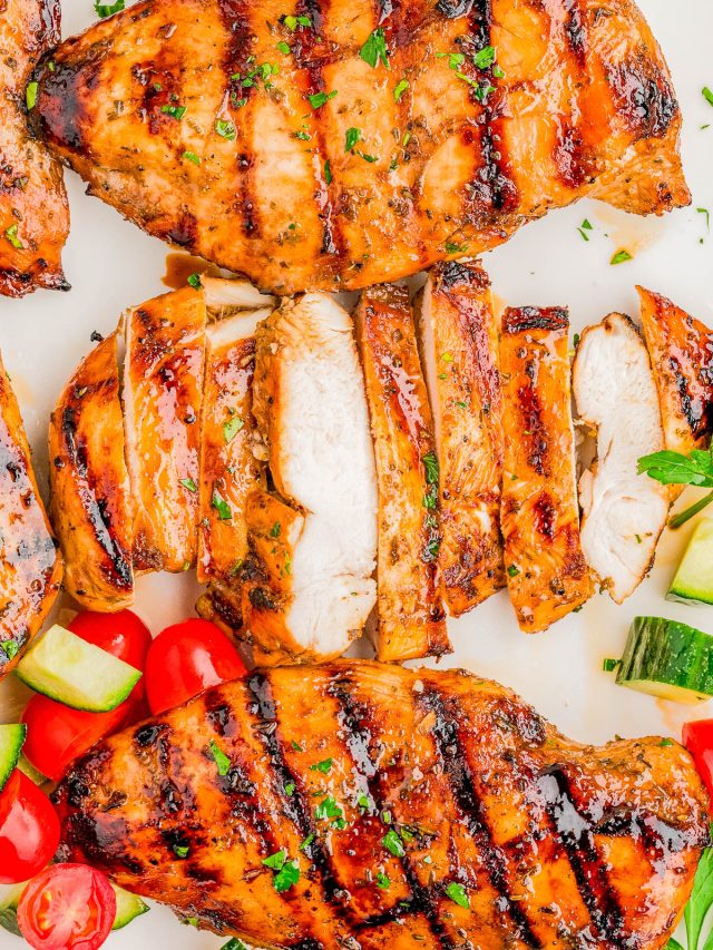 Grilled chicken breasts, some sliced, garnished with herbs and served with cherry tomatoes and cucumber pieces on a white surface.