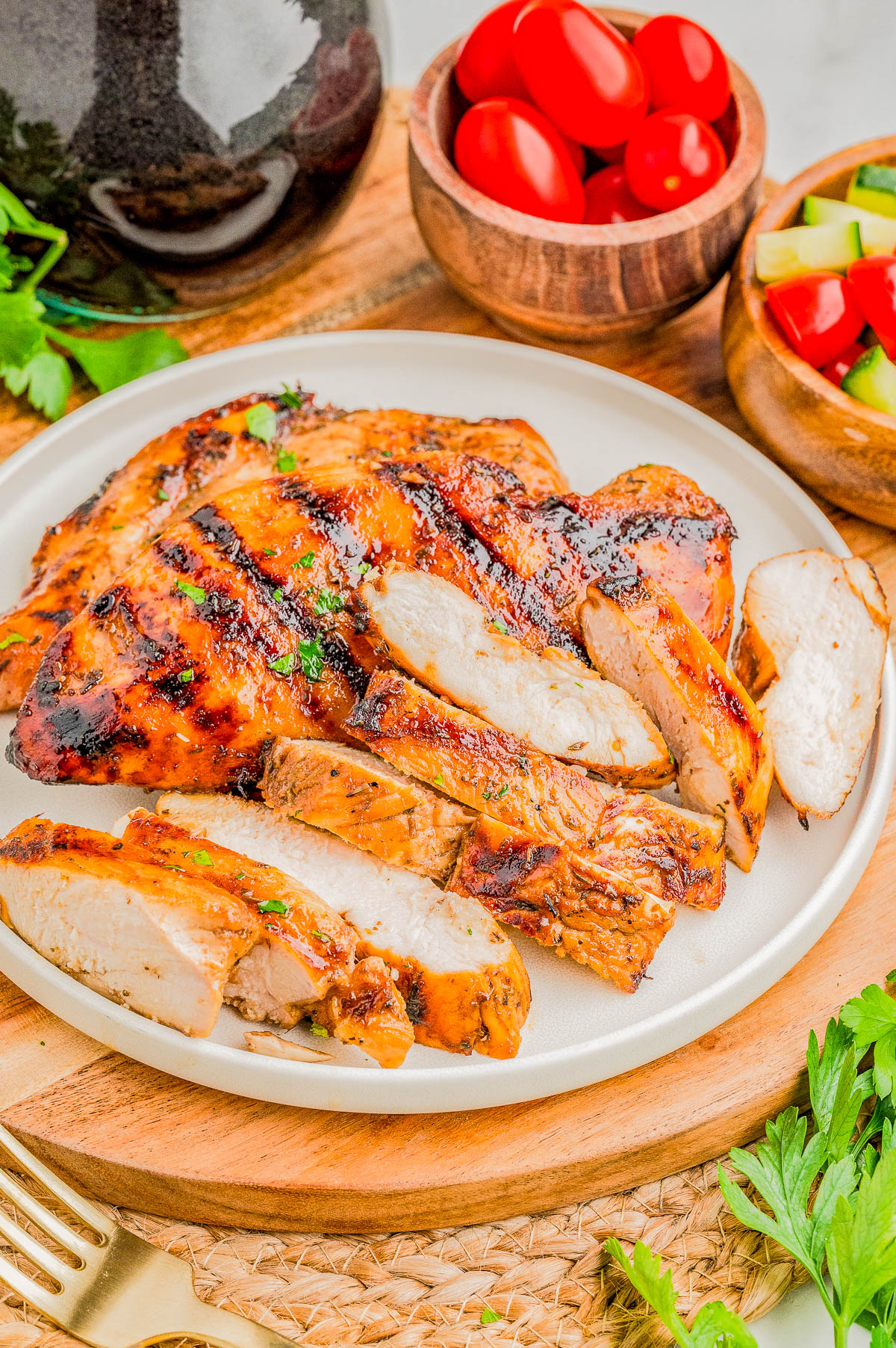 Sliced grilled chicken breast on a white plate, served with fresh parsley, cherry tomatoes in a wooden bowl, and a bowl of chopped vegetables.