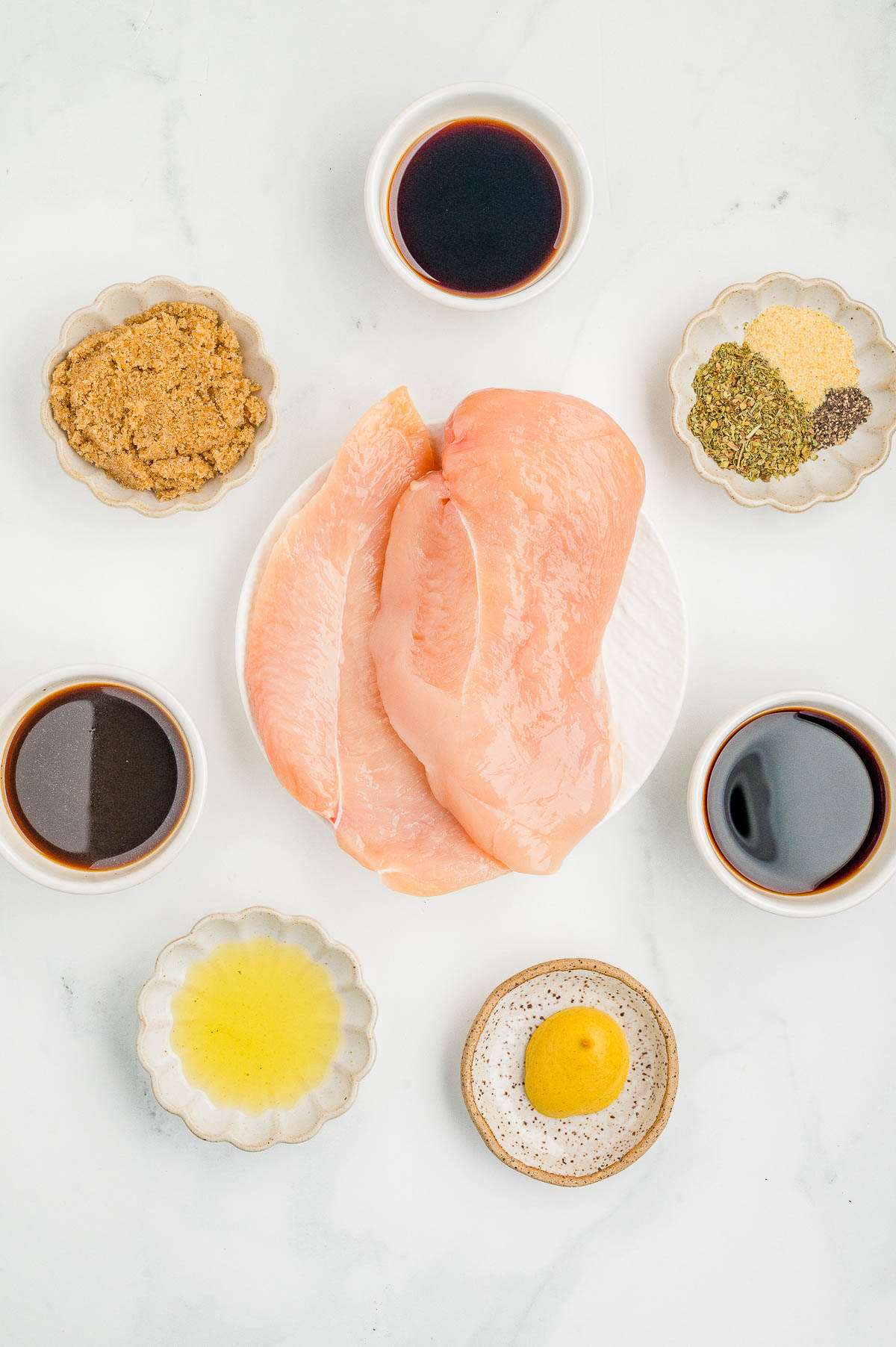 Raw chicken breasts on a plate surrounded by small bowls containing soy sauce, Worcestershire sauce, spices, brown sugar, olive oil, and mustard, arranged on a white surface.
